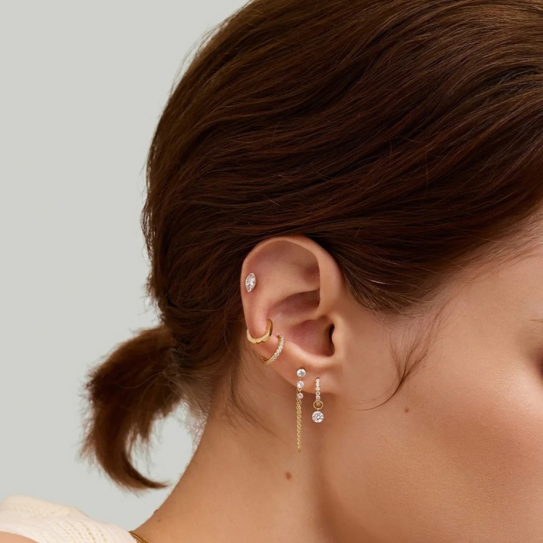 A lifestyle image of a woman's ear showing the 4mm diamond charm drop hanging on a diamond huggie, along with a number of other earrings worn in her lobes and cartilage.   