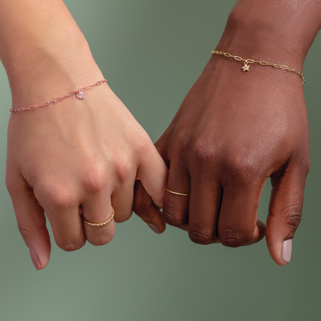 A close-up image of two women's hands and wrists, with pinky fingers interlocked, featuring their 14k gold forever linked paperclip chain with petite charm dangles from the Forever Linked collection | Peterson MADE