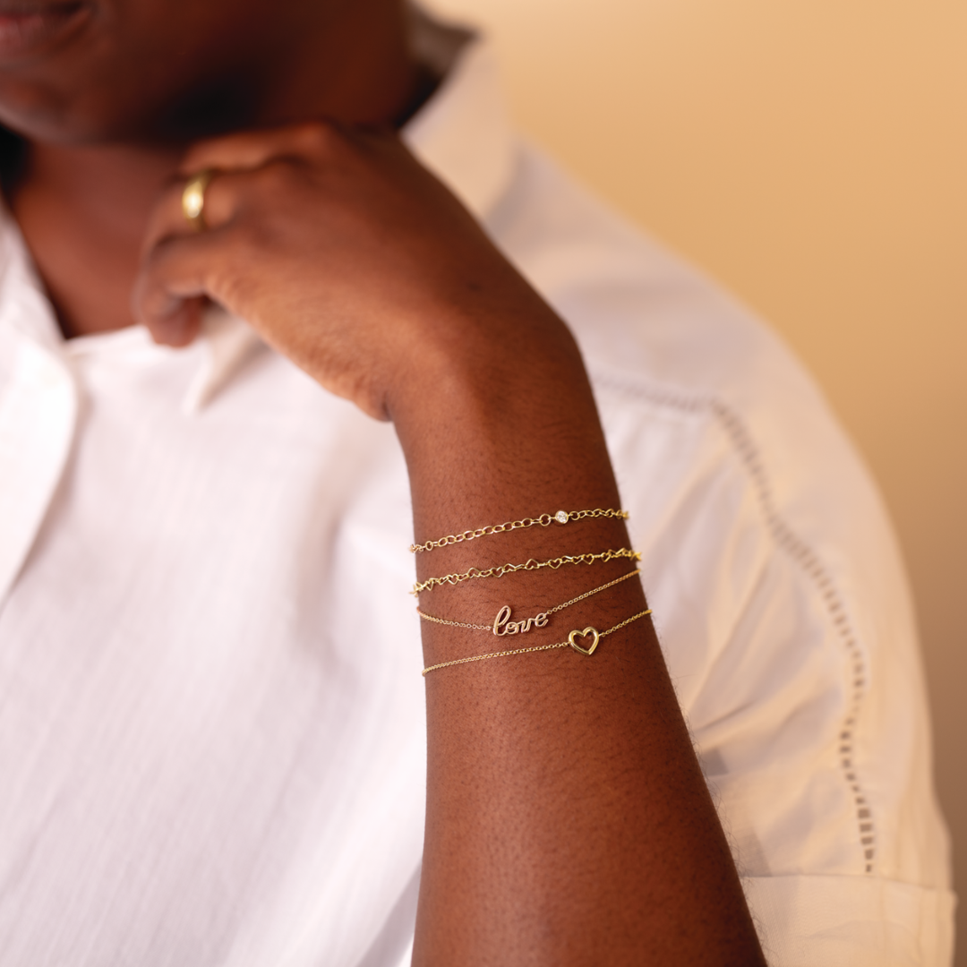 A woman wearing a white shirt with a neutral backdrop with the image focused on her wrist, featuring a collection of 4 14k gold forever linked permanent bracelets.  1 has no charm, and the remaining 3 features a heart link, a script love link, and a diamond gemstone bezel link. 