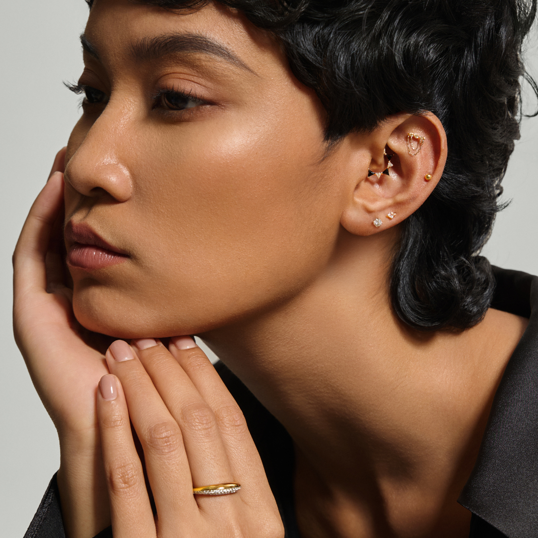 Close-up of a person with short hair, wearing earrings and a ring, against a neutral background. Featuring her ear stack of comfortable piercing jewelry. 