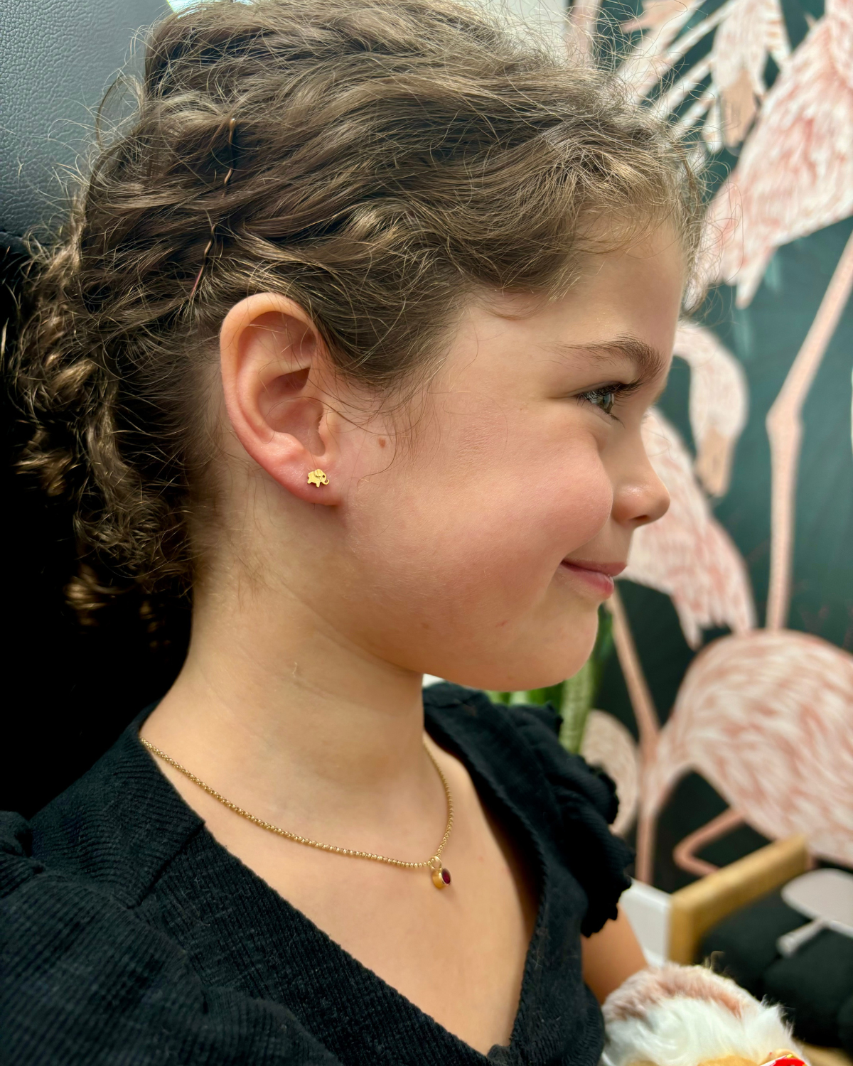 A young girl with curly brown hair is shown just after her piercing appointment, wearing a Titanium Elephant Threadless Top in her lobe and a delicate gold necklace with a tiny red birthstone.  She is wearing a black top, and a patterned wallpaper featuring pink flamingos is visible in the background.