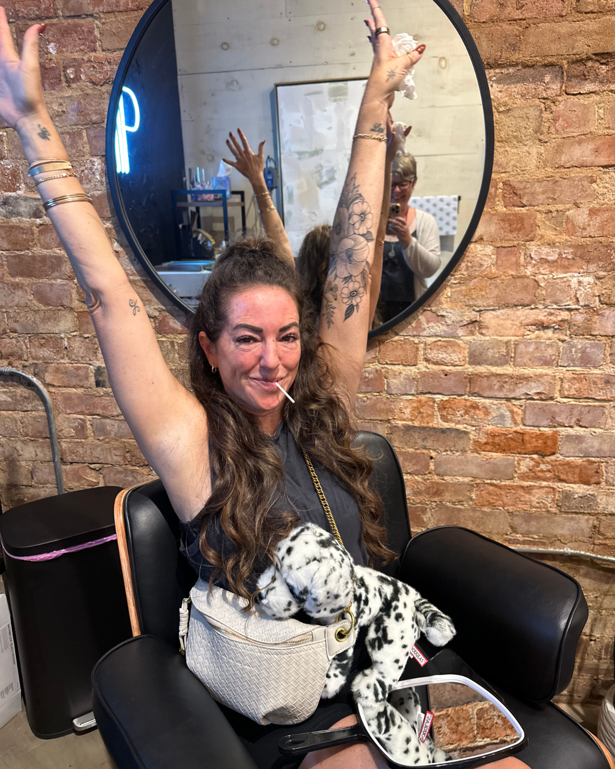 A woman sitting in the piercing chair with her arms raised in victory and excitement having just been pierced by Peterson MADE.  She's sitting in a piercing / salon chair with a brick wall behind her.